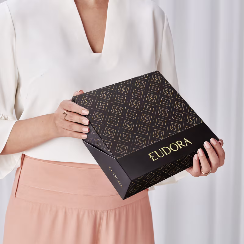A caixa preta com detalhes dourados apresenta o logotipo da marca Eudora. A caixa possui um padrão geométrico repetido em sua superfície. Uma mulher está segurando a caixa.