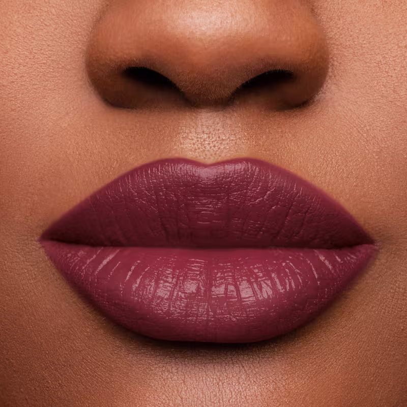 Foto com foco na boca da modelo com o produto Batom. Acabamento brilhante e uma cor predominante de vermelho escuro. A superfície do batom é lisa e brilhante, com uma textura cremosa.