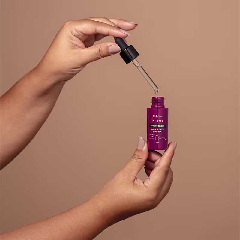 O Potencializador de Nutrição da linha Siàge Pro Cronology está sendo segurado por uma mão, aberto. O frasco tem um conta-gotas preto na tampa. Parece ter uma consistência líquida e é apresentado em um ambiente com fundo bege.