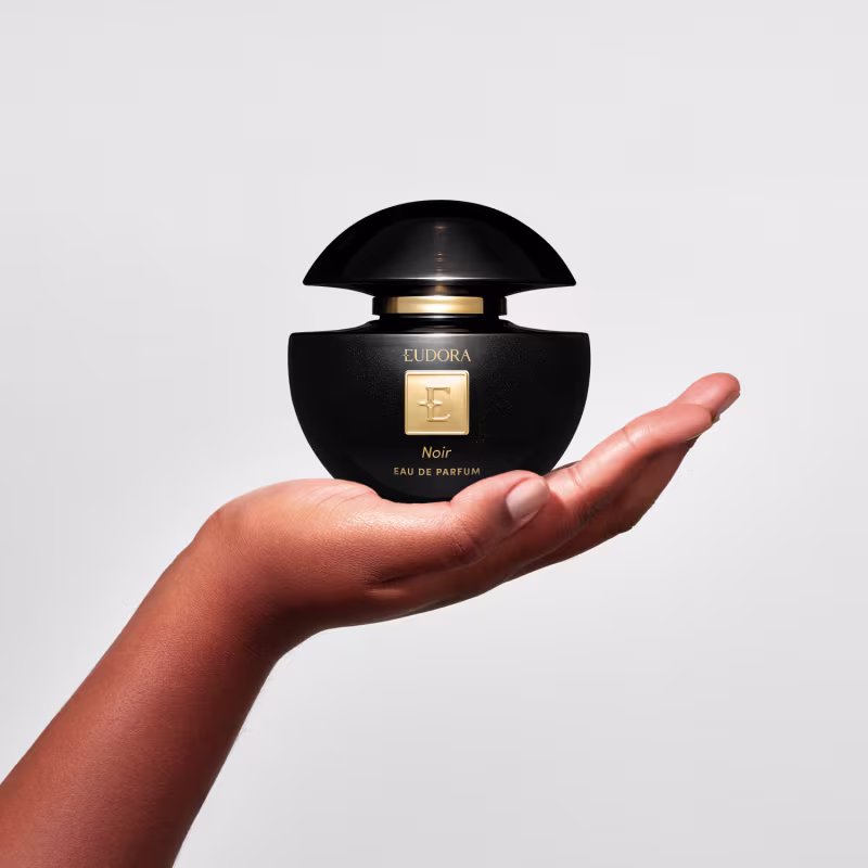 Mão feminina segurando um frasco de Eudora Noir Eau de Parfum preto e dourado da marca Eudora, em um ambiente claro e minimalista, sugerindo um contexto de beleza e sofisticação.