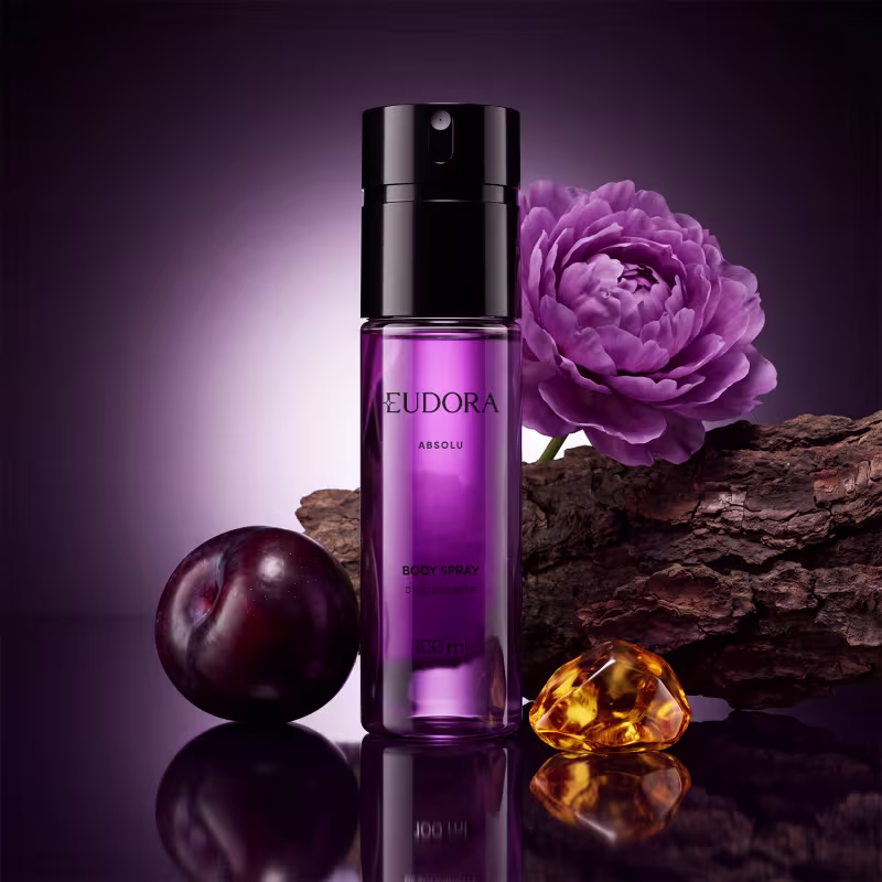 Frasco transparente, com líquido roxo claro e tampa preta, do item de perfumaria Eudora Absolu. Com uma flor de peônia roxa e ameixa preta ao redor, além de casca de Sândalo e pedra de âmbar dourado, indicando os ingredientes olfativos.
