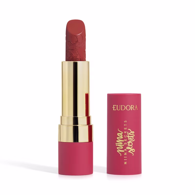 Embalagem de um batom aberto, com corpo dourado e tampa e base rosa, escrito "Eudora Niina Secrets". A cor do batom é vermelha e tem um relevo exclusivo em padrão floral na bala. Produto de beleza com design elegante e sofisticado.
