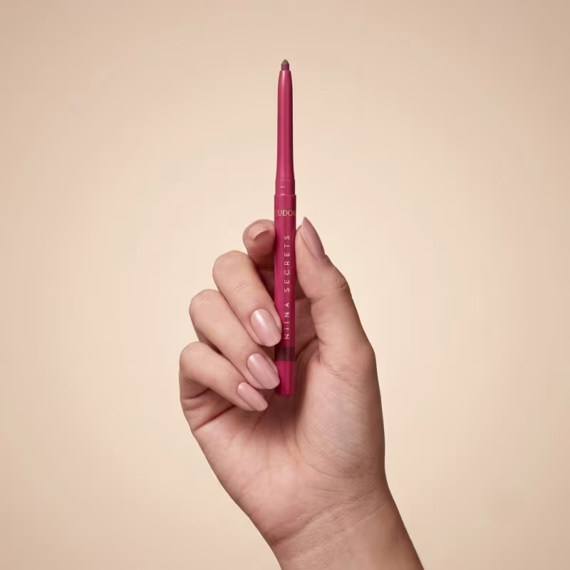 Lápis labial rosa da marca Niina Secrets sendo segurado por mão feminina com unhas curtas e esmaltadas em fundo bege claro.