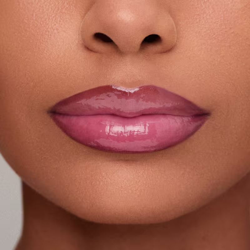 Imagem de close-up de lábios femininos com lapiseira labial e gloss amarronzado brilhante, em fundo claro e suave. 
