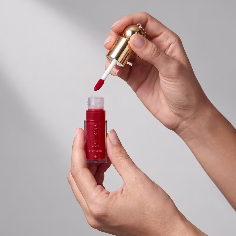 Mãos segurando o batom líquido aberto, com aplicador. O item está sobre um fundo cinza claro. A cor do produto é em tom de vermelho.
