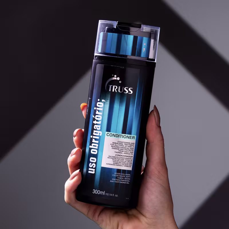 A imagem mostra uma mão segurando um frasco de condicionador para cabelos da marca Truss. O frasco é azul escuro e preto. Na parte frontal, está escrito "Uso Obrigatório" em letras brancas.