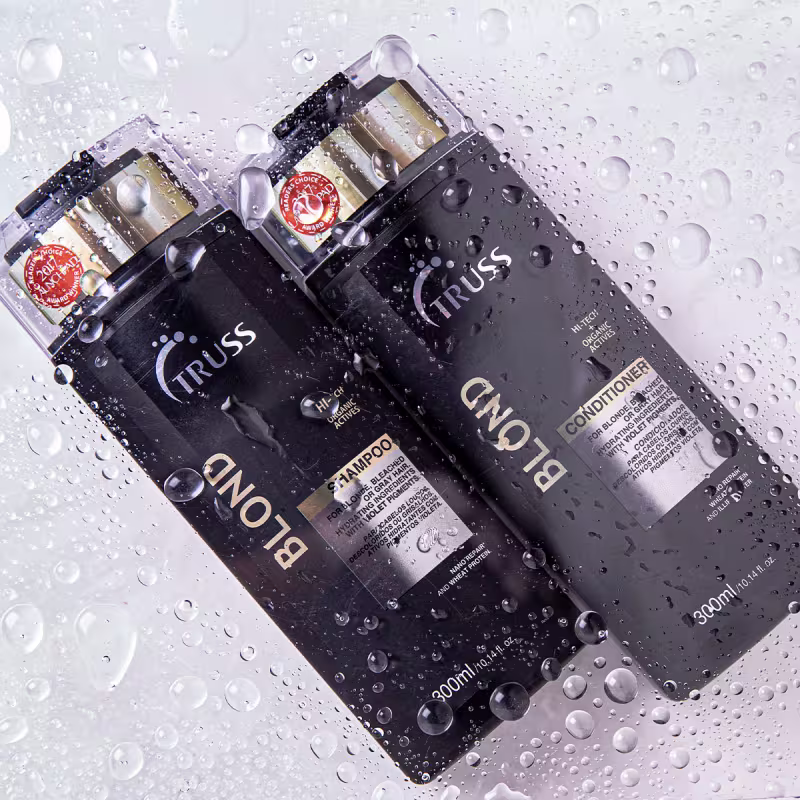 A imagem mostra dois frascos de shampoo e condicionador da marca Truss, da linha Blond. Os frascos são de cor preta e estão molhados, com várias gotas d'água condensadas em sua superfície, criando um efeito de fundo desfocado e brilhante.