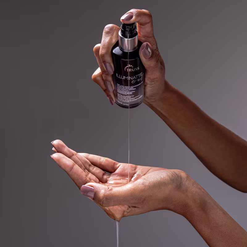 A imagem mostra um par de mãos segurando um frasco preto com o rótulo "Truss Illuminatte". Uma das mãos está segurando o frasco enquanto a outra está aberta embaixo, como se estivesse aplicando ou recebendo o produto do frasco.