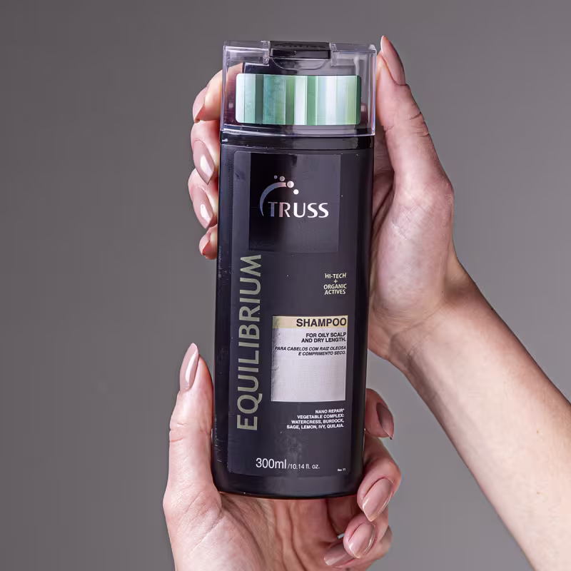 A imagem mostra uma mão segurando um frasco de shampoo da marca Truss, da linha Equilibrium. O frasco é preto e tem uma tampa transparente. O rótulo indica que é um shampoo para couro cabeludo oleoso e cabelos secos.