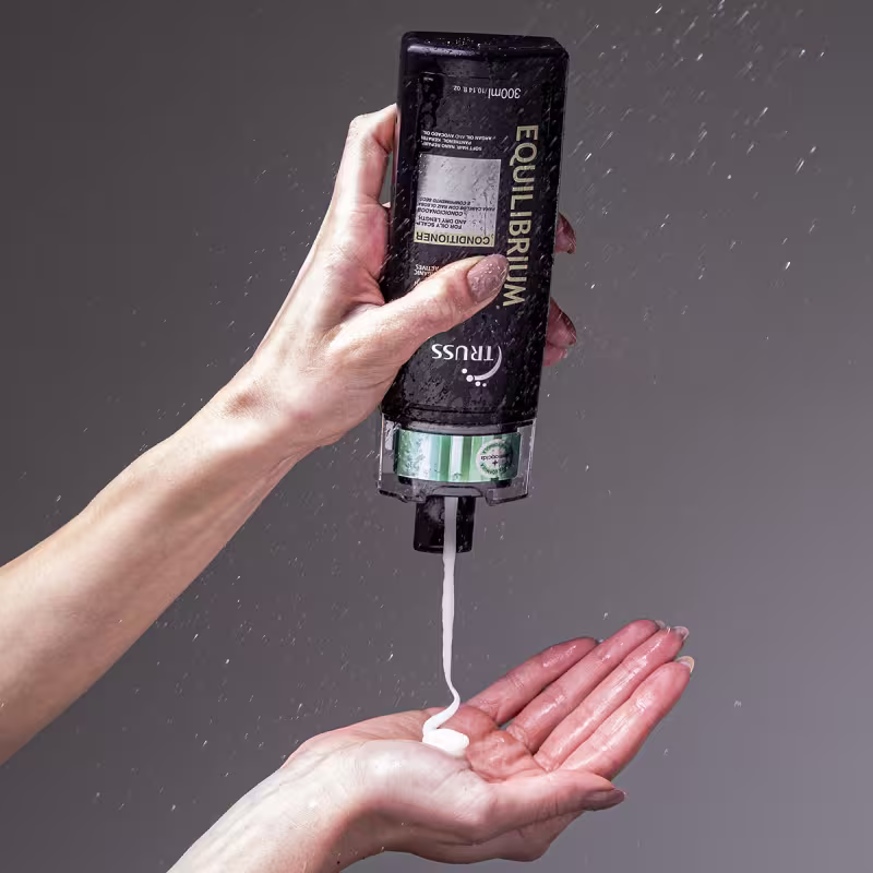 A imagem mostra uma mão segurando um frasco de condicionador da marca Truss, linha Equilibrium. O frasco está molhado, aparentemente sendo usado no chuveiro ou banheiro. Também é possível ver o seu conteúdo, que é cremoso e branco.