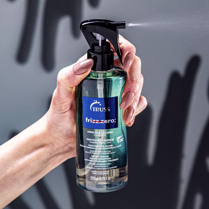 A imagem mostra uma mão segurando um frasco de um produto capilar da marca Truss chamado "frizz.zero". É uma máscara líquida para proteção térmica e controle do frizz.