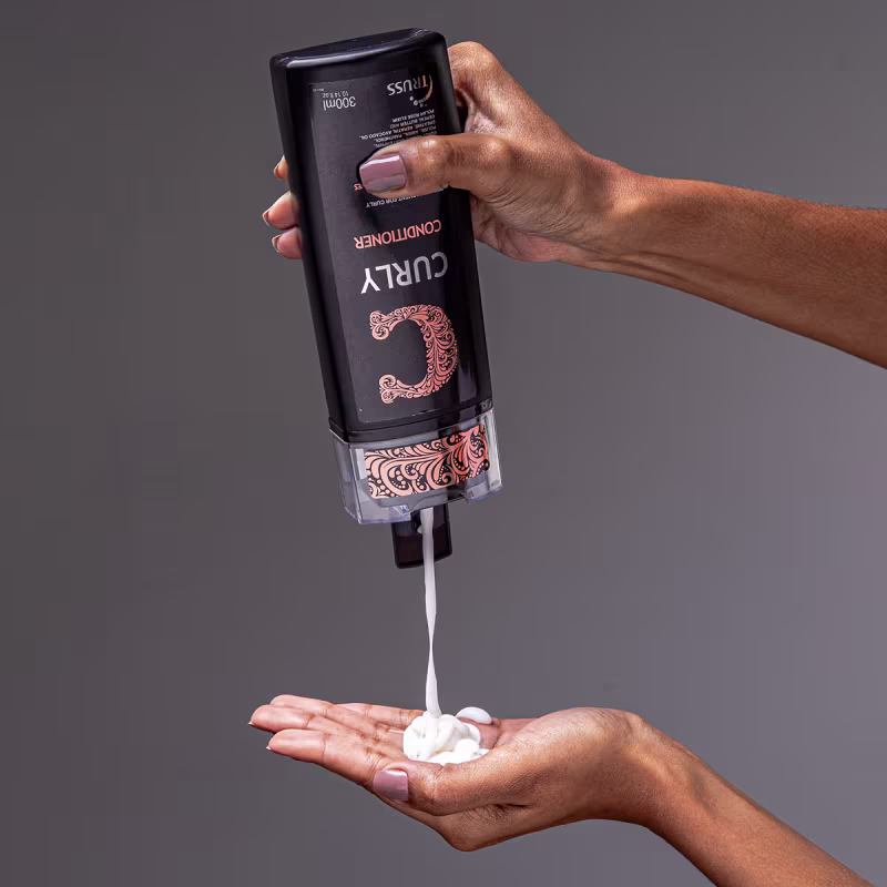 A imagem mostra o Condicionador TRUSS Curly. O líquido está sendo despejado do frasco em uma mão feminina, mostrando a espessura do produto.