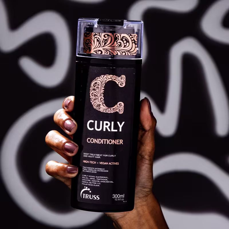 A imagem mostra uma mão feminina com unhas esmaltadas de rosa, segurando o Condicionador Curly da marca TRUSS, uma embalagem preta com detalhes em arabescos e tom laranja.