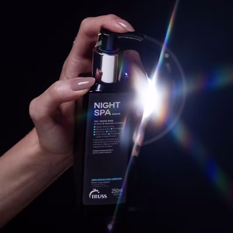 A imagem tem fundo preto e duas mãos femininas segurando o produto TRUSS Night Spa, frasco preto com tampa metálica. Nela, podemos ver uma mão segurando a embalagem; enquanto a outra segura o conteúdo despejado.