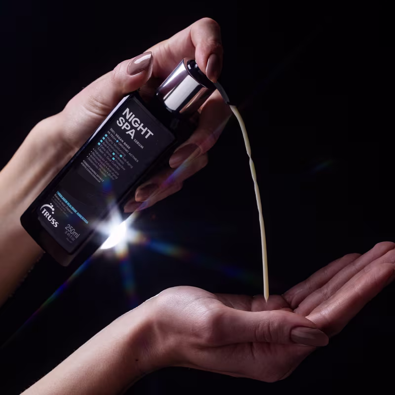 A imagem tem fundo preto e duas mãos femininas segurando o produto TRUSS Night Spa, frasco preto com tampa metálica. Nela, podemos ver uma mão segurando a embalagem; enquanto a outra segura o conteúdo despejado.