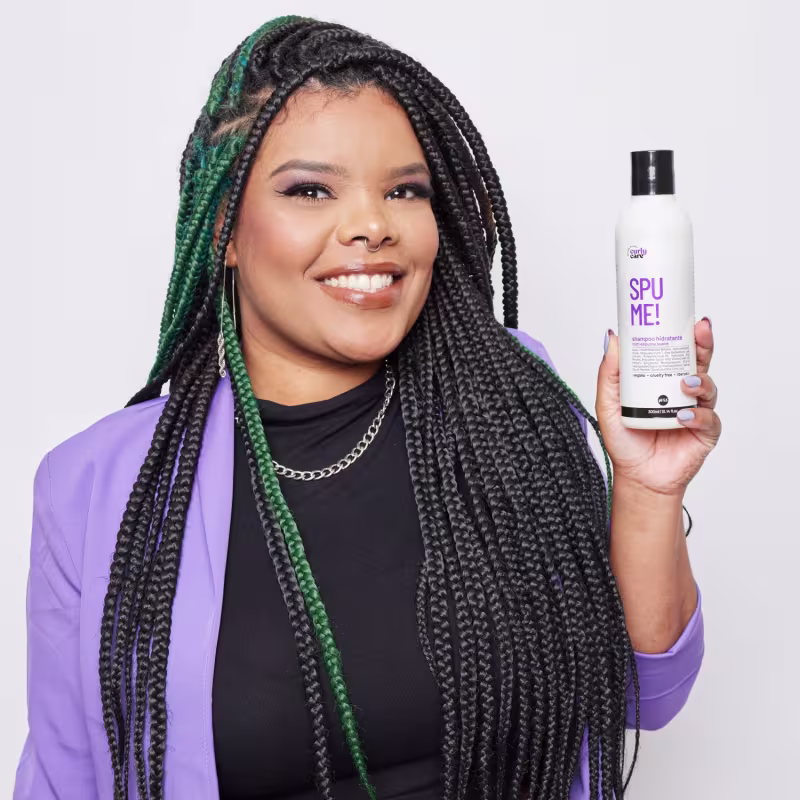 O produto mostrado é um frasco plástico branco para cabelos cacheados chamado "Spu Me!". O frasco tem uma tampa preta e o rótulo é predominantemente roxo com o nome do produto em letras grandes.