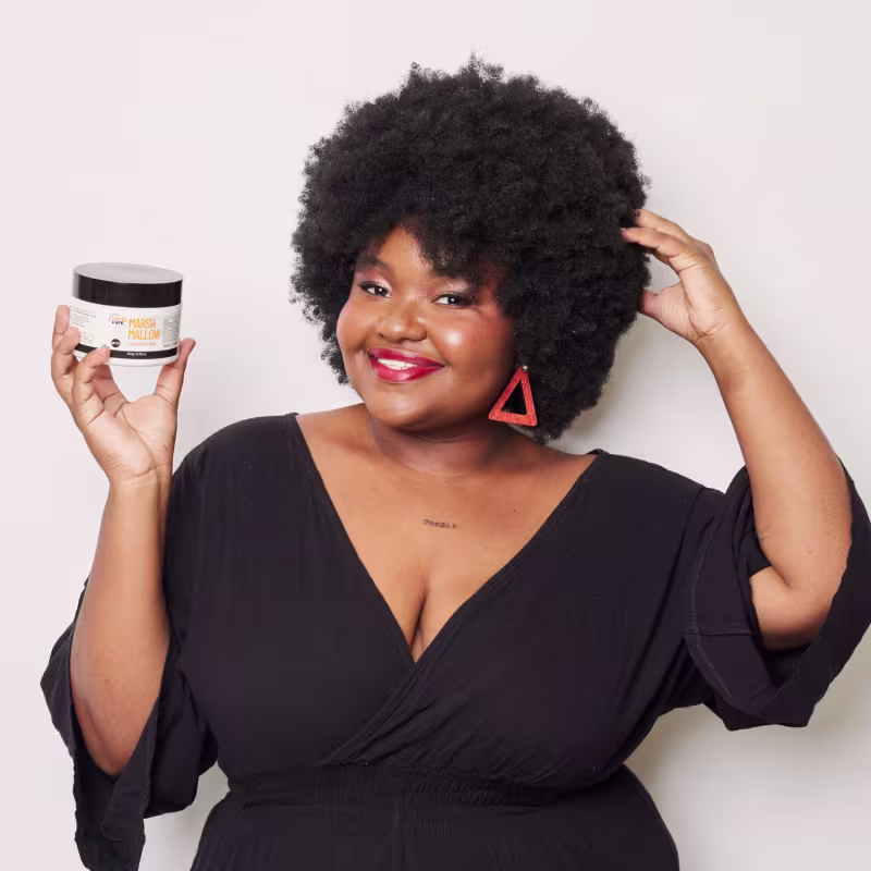 É um pote de máscara capilar da linha Curly Care. O pote é branco com detalhes em preto. Na parte frontal, está escrito "MARSH MALLOW máscara HNR" em letras grandes e laranjas.