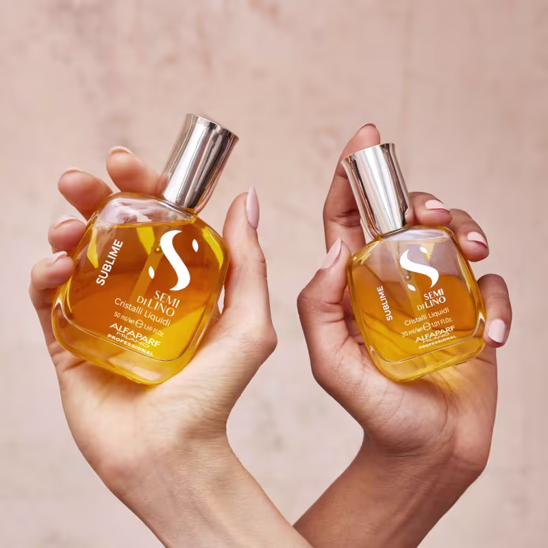 Dois frascos de perfume da marca Alfaparf Milano. Os frascos são de vidro âmbar com tampas metálicas prateadas. Nas embalagens, há o logotipo da marca e o nome do produto "Semi di Lino Cristalli Liquidi" impresso, além das informações "50 ml/1.69 fl.oz.".