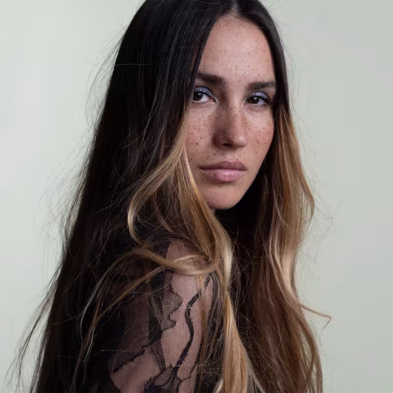 Esta imagem mostra uma mulher jovem com cabelos longos e ondulados em tons de castanho escuro e loiro claro. A expressão da mulher é séria e contemplativa, com olhos azuis penetrantes e lábios naturais.