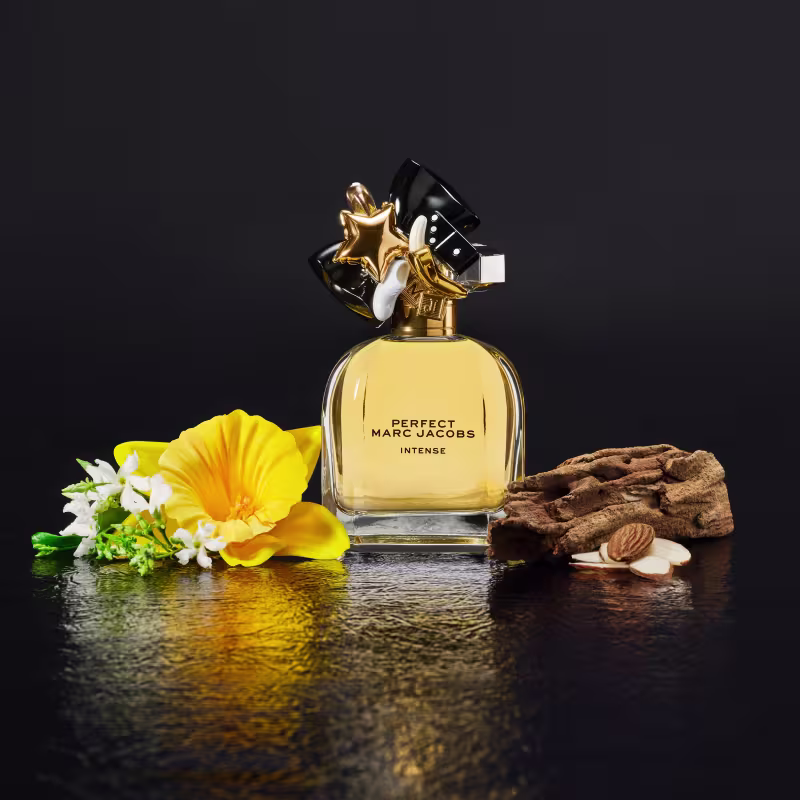 Frasco de perfume dourado e preto da linha "Perfect" da marca Marc Jacobs, com detalhes em estrela e fita preta na tampa. Rótulo com o nome do produto "Perfect Marc Jacobs Intense" em letras pretas.