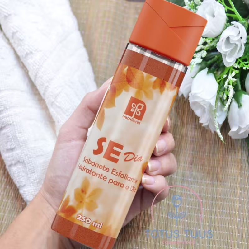 Sabonete esfoliante hidratante para o dia da marca Natuflores. Embalagem em frasco cilíndrico transparente de 250ml com tampa laranja. Rótulo laranja com detalhes em branco e amarelo, exibindo o nome do produto "SE Dia".
