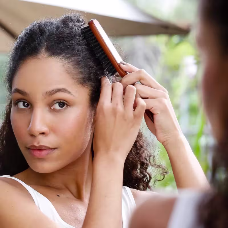 Esta imagem mostra uma mulher de cabelos cacheados escuros usando a escova de cabelo de madeira. Ela está penteando seus cabelos com cuidado. A mulher veste uma regata branca e tem um olhar concentrado enquanto arruma o cabelo.