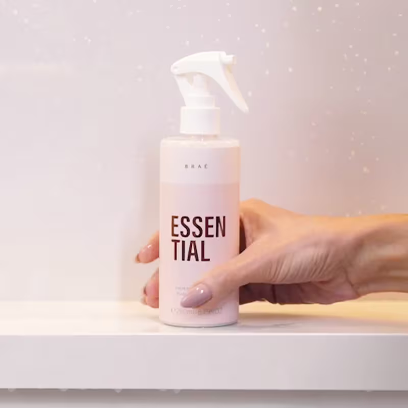 Produto de cuidados pessoais da marca Braé. Frasco plástico spray transparente com líquido rosa claro. Rótulo com o texto "ESSENTIAL" em letras maiúsculas pretas. Mão segurando o frasco, com unhas pintadas de lilás.