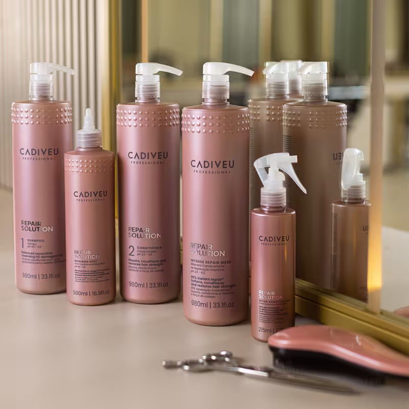 Os produtos da linha estão em dispostos em uma penteadeira, à frente de um espelho. Ao redor há uma tesoura e uma escova de cabelo.