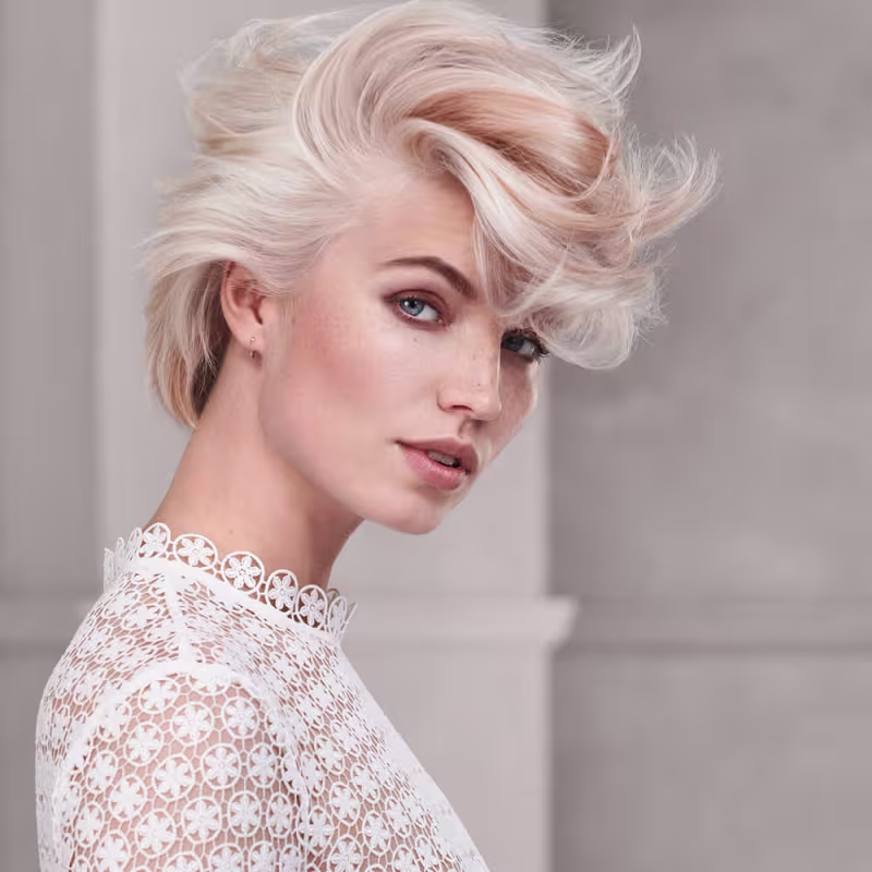 Esta imagem apresenta uma modelo feminina com cabelos platinados e estilo despojado. Ela está usando uma blusa rendada branca com detalhes em formato de flores vazadas.