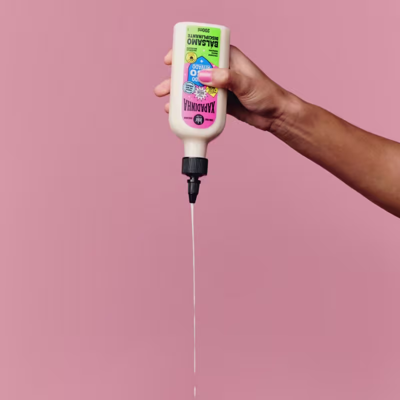 A imagem mostra uma mão derramando o bálsamo. É possível ver um líquido branco formando um longo fio ao sair da tampa.