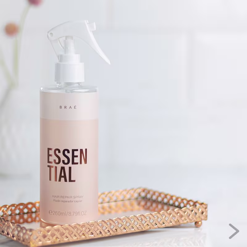 Produto de reparação capilar em spray da marca Brae. Embalagem plástica transparente e rosa claro com o nome "Essential" em letras marrons. Frasco de spray com aplicador de bomba.