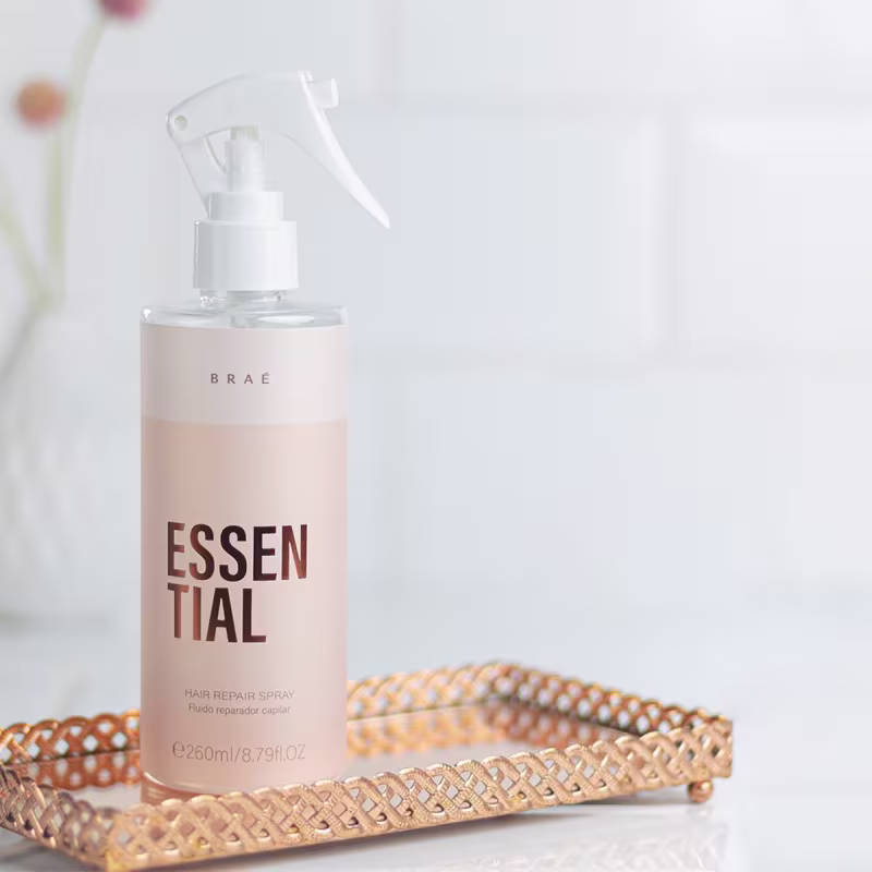 Produto de reparação capilar em spray da marca Brae. Embalagem rosa claro. Rótulo com o nome "Essential". Frasco plástico transparente com bomba pulverizadora branca.