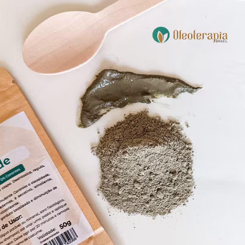 Imagem mostra uma superfície branca e sobre ela está: a embalagem da argila verde, uma colher de madeira pequena, uma amostra da argila úmida pronta e uma porção pequena da argila em pó.