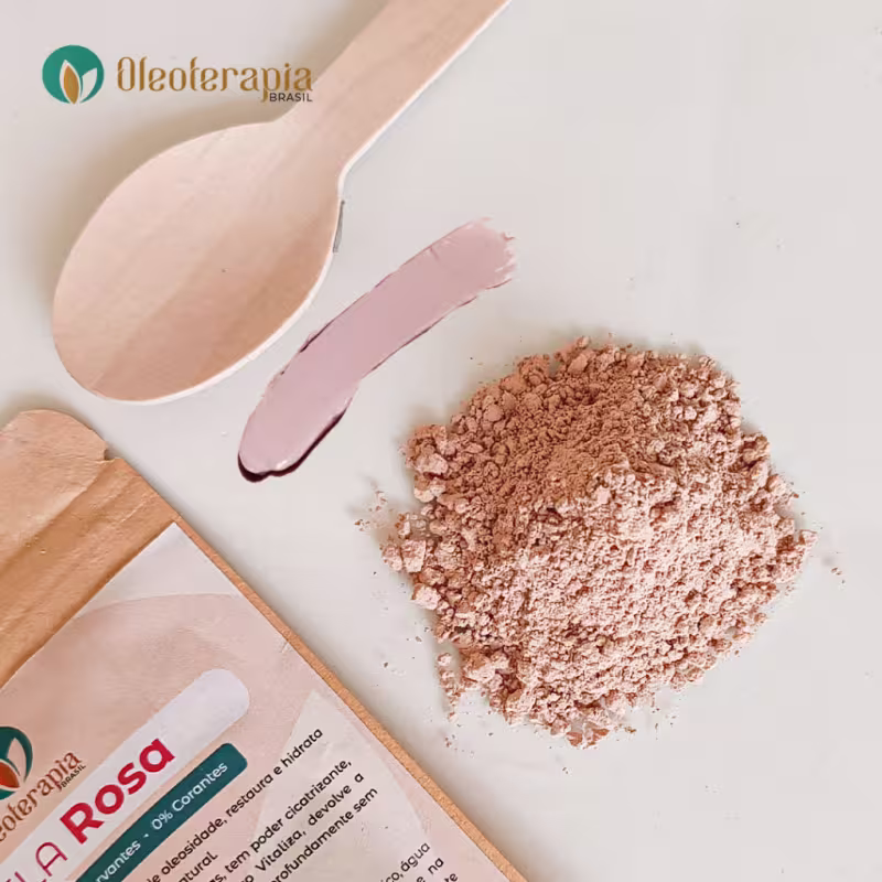 A imagem mostra uma superfície branca e sobre ela está: a embalagem da argila rosa, uma colher de madeira pequena, uma amostra da argila úmida pronta e uma porção pequena da argila em pó.