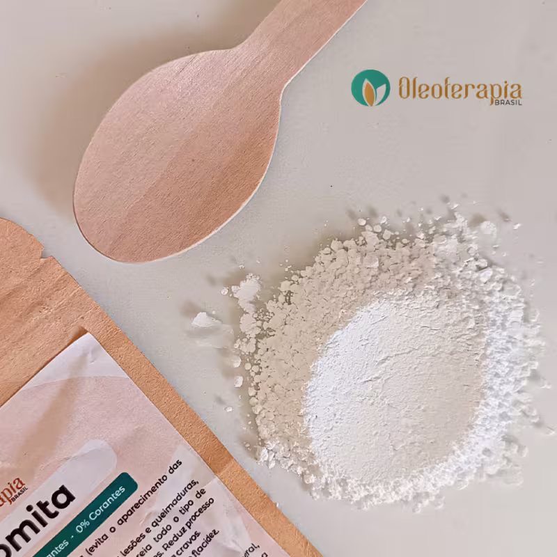 Imagem mostra uma superfície branca e sobre ela está: a embalagem da dolomita, uma colher de madeira pequena, uma porção pequena da dolomita em pó, sua cor é branca.