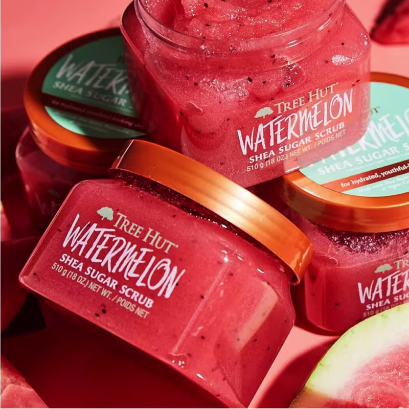 Produto de esfoliação corporal Tree Hut Watermelon Shea Sugar Scrub. Embalagem transparente com tampa laranja brilhante. O frasco contém um esfoliante de açúcar de cor rosa vibrante com pequenas sementes escuras visíveis.