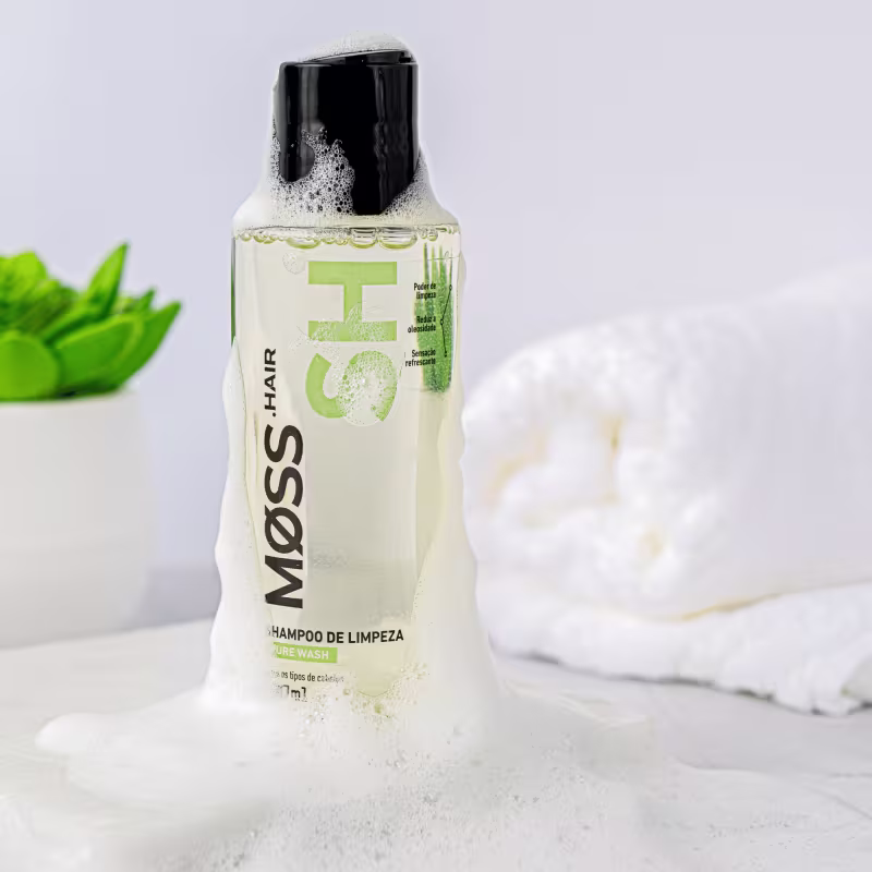 A imagem mostra o frasco do shampoo Pure Wash em uma superfície branca coberto de espuma. Ao fundo tem uma toalha branca e um vaso branco com uma planta de folhas verdes.