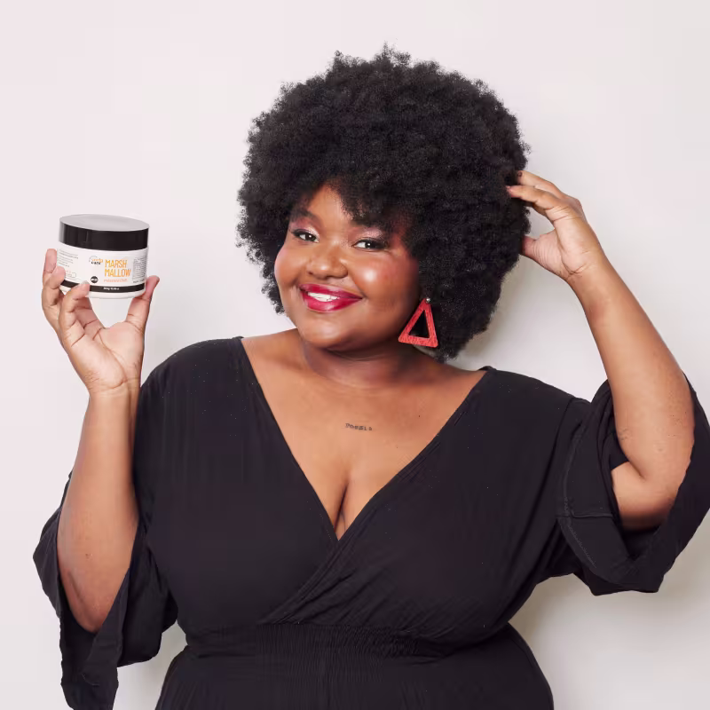 O produto mostrado é um creme para cabelos cacheados em um pote branco com tampa preta. A embalagem tem o rótulo "Marsh Mallow" escrito nela. A cor predominante é branca com detalhes em preto.