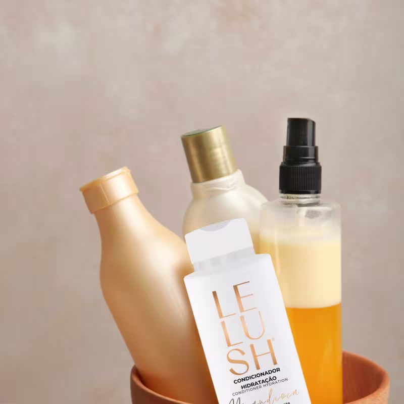 A imagem mostra três frascos de produtos cosméticos nas cores dourada e amarelo juntos em um recipiente. Entre eles está o frasco do condicionador "Lelush".