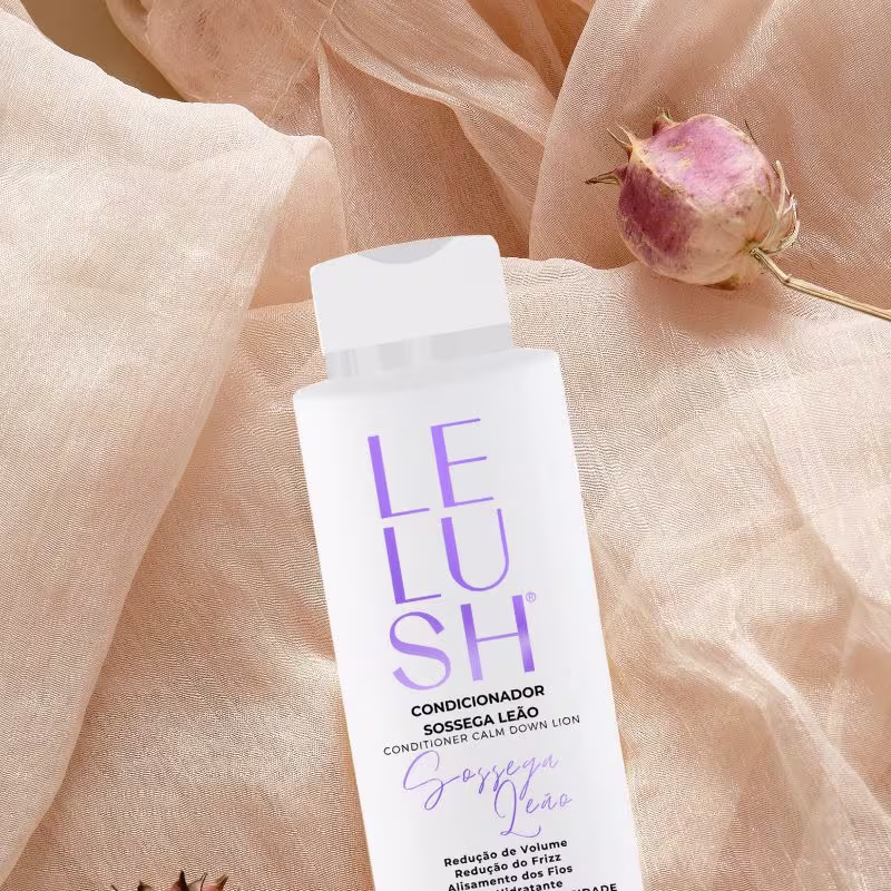 A imagem mostra um frasco de condicionador branco com o nome da linha escrito em letras roxas. A composição também inclui um tecido bege delicado e um botão de rosa rosada, criando uma atmosfera suave e natural.