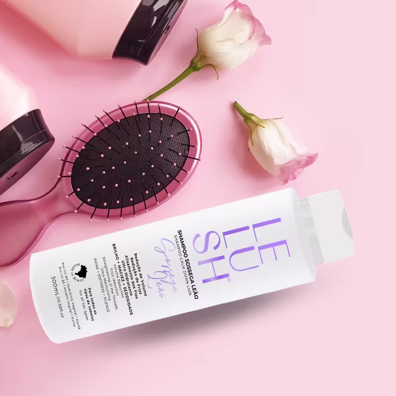 A imagem mostra uma escova de cabelo redonda com cerdas pretas e detalhes rosa, duas flores brancas delicadas e um frasco branco com o shampoo. Os itens estão dispostos sobre um fundo rosa claro, criando uma atmosfera delicada.