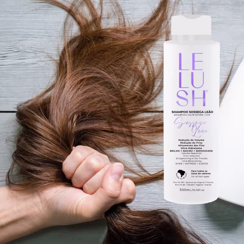 A imagem mostra uma mão segurando delicadamente alguns fios de cabelo longos e ondulados de cor castanha. Ao lado, há um frasco branco com o shampoo da linha Lelush Sossega Leão.