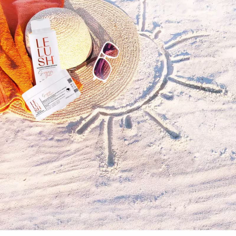 Temos o pote branco de máscara capilar junto com o shampoo, ao lado, há um chapéu de palha tipicamente usado na praia, óculos de sol espelhados e uma toalha laranja. No fundo, desenhado na areia, há um grande sol estilizado com diversos raios ao redor.