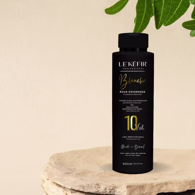 A imagem mostra a embalagem preta da água oxigenada posicionada sobre uma pedra clara, em um fundo bege, com folhas verdes decorando a cena, transmitindo uma atmosfera natural para esse cosmético capilar.