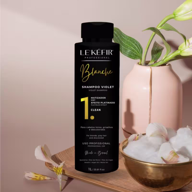 A imagem mostra a embalagem do shampoo posicionada ao lado de uma vasilha dourada com plantas e flores brancas, criando um cenário natural e elegante para apresentar o produto. As flores parecem ser orquídeas, com suas folhas verdes e hastes.