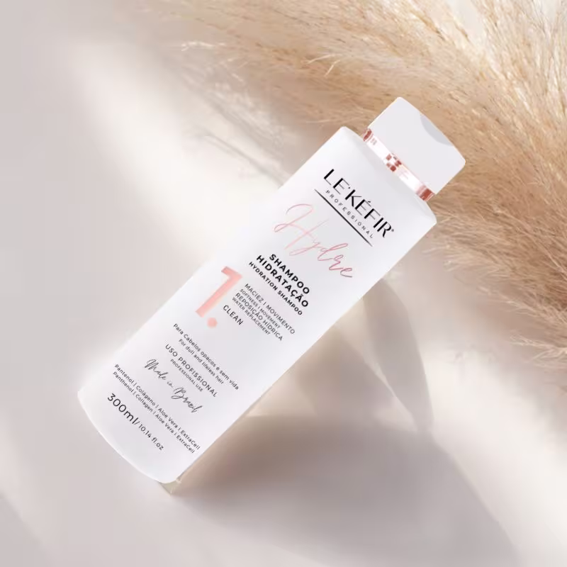 A imagem tem um fundo claro com o frasco do shampoo deitado em destaque na parte central da imagem. No canto direito dando um toque sutil ao cenário há um pedacinho de haste de capim dos pampas.