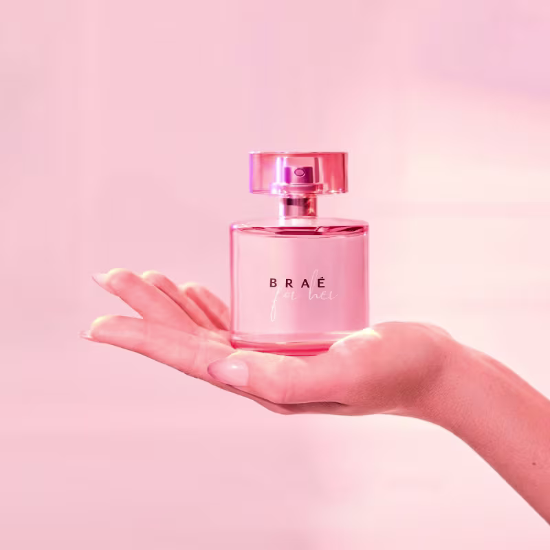 Perfume em frasco de vidro transparente na cor rosa. O frasco tem um formato retangular com cantos arredondados e uma tampa rosa brilhante. O perfume é segurado por uma mão feminina. O fundo é  na mesma tonalidade rosa clara