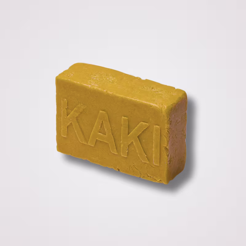 Barra de sabonete retangular de cor amarelo-mostarda. A palavra "KAKI" está gravada em relevo maiúsculo na superfície do sabonete. A textura aparenta ser levemente áspera e rústica. O produto tem uma forma sólida e compacta, típica de sabonetes artesanais