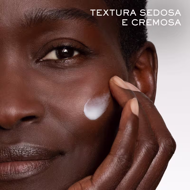A imagem mostra um close-up de uma mulher negra aplicando um creme ou produto de beleza cremoso na área ao redor do olho. O produto tem uma textura sedosa e cremosa, conforme indicado pelo texto "Textura Sedosa e Cremosa".