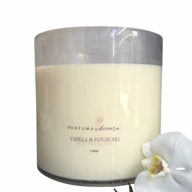 Vela aromática em frasco de vidro cilíndrico transparente. O frasco é preenchido com cera bege claro. Rótulo na parte frontal indica "Perfuma & Decora, Vanilla & Patchouli" e o peso de 4,5kg. Uma pequena flor branca de orquídea está ao lado do frasco.